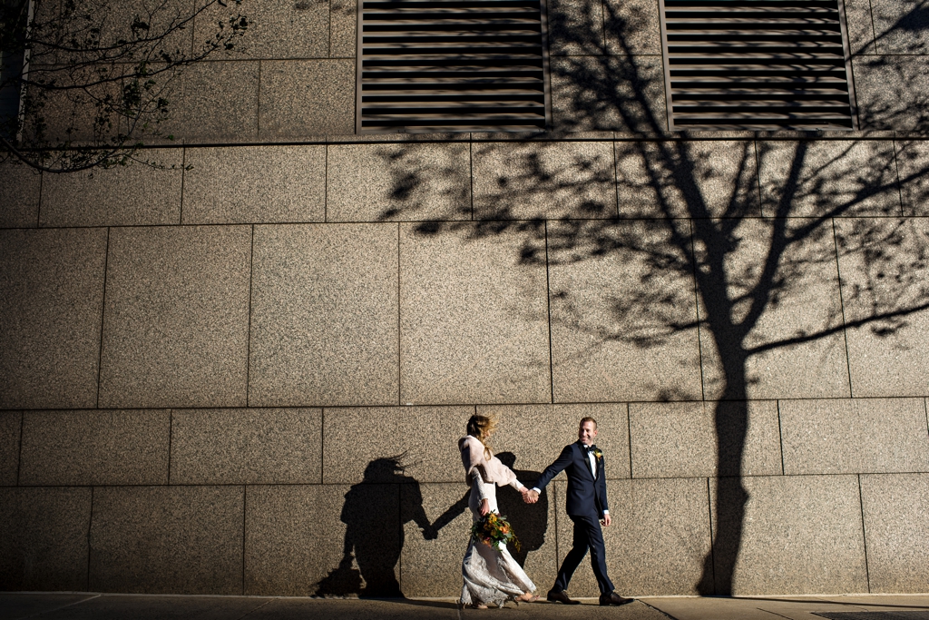 A groom walks with his bride down the Philly streets with a shadow of a tree against the wall.